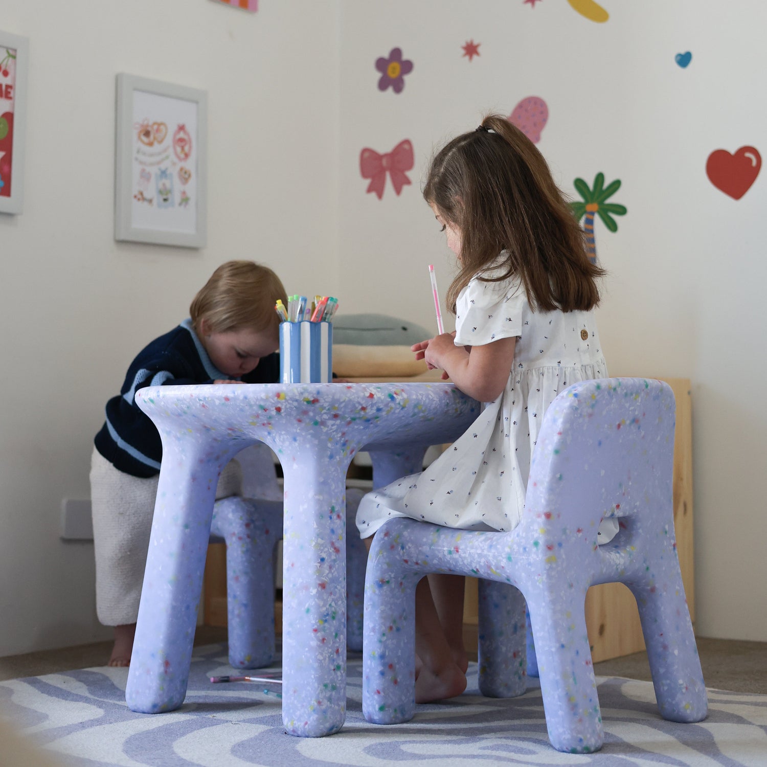 Two children sitting at a small table with colorful wall stickers in a child-friendly room.