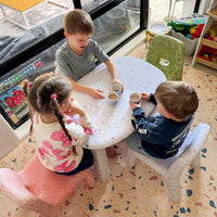 Three children sitting around a small round table in a room with large windows.